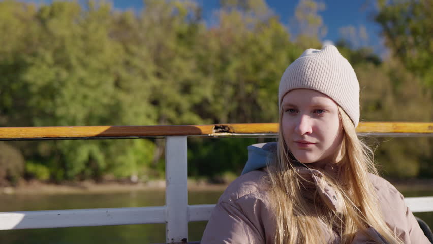 Teenage girl passenger sitting on boat near Lake Ontario shore, young woman in warm clothes enjoying calm travel moment with thoughtful expression and autumn nature background.
