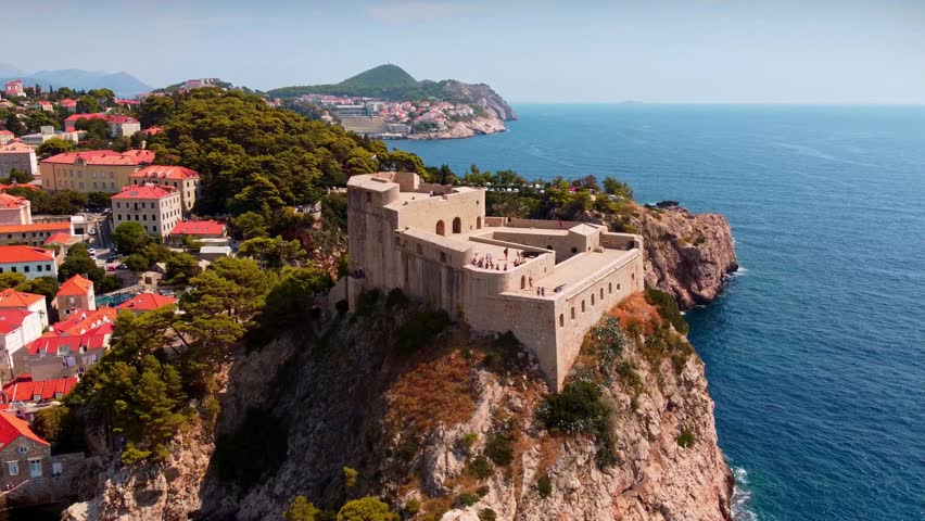 Aerial drone view of Fort Lovrijenac perched on a rocky cliff above the Adriatic Sea in Dubrovnik, Croatia, with historic stone walls, red rooftops, and lush coastal scenery.