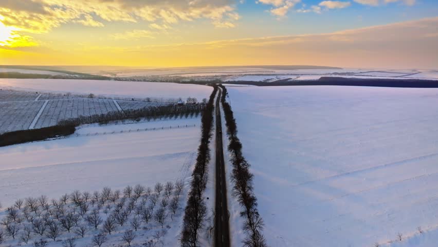 Drone footage soaring above snowy winter road at sunset, trees lining asphalt path leading to village.