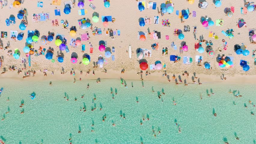 Top down aerial drone view of a crowded beach with colorful umbrellas and people relaxing. Turquoise crystal clear sea water and white sand during a bright sunny summer vacation.
