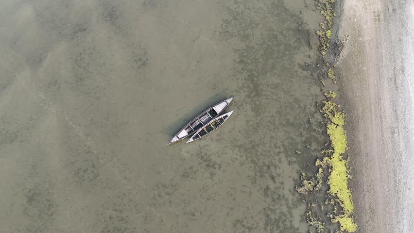 Serene aerial top down perspective showing two traditional wooden canoes floating peacefully on the calm, murky surface of a shallow river next to a sandy and mossy shore