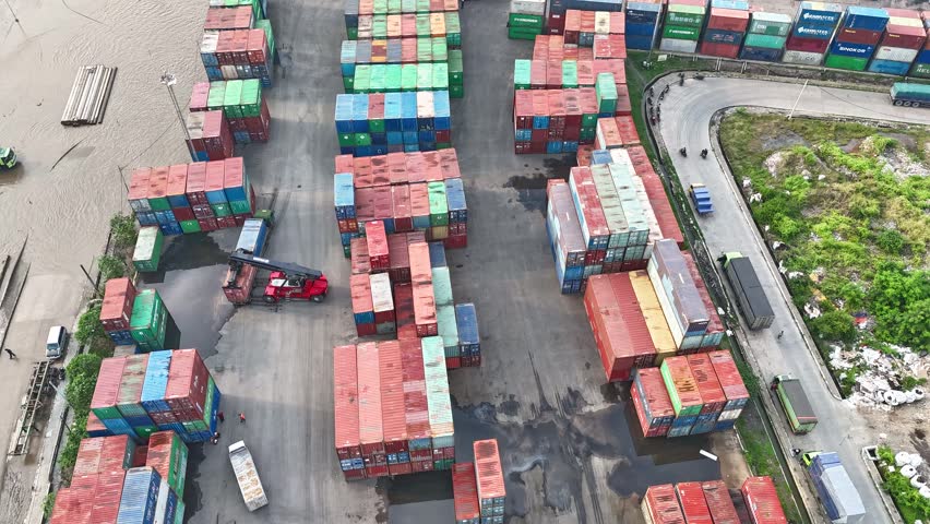 High-angle aerial drone shot of a busy logistics terminal featuring a reach stacker crane moving industrial shipping containers in a large storage yard for global import export trade operations.
