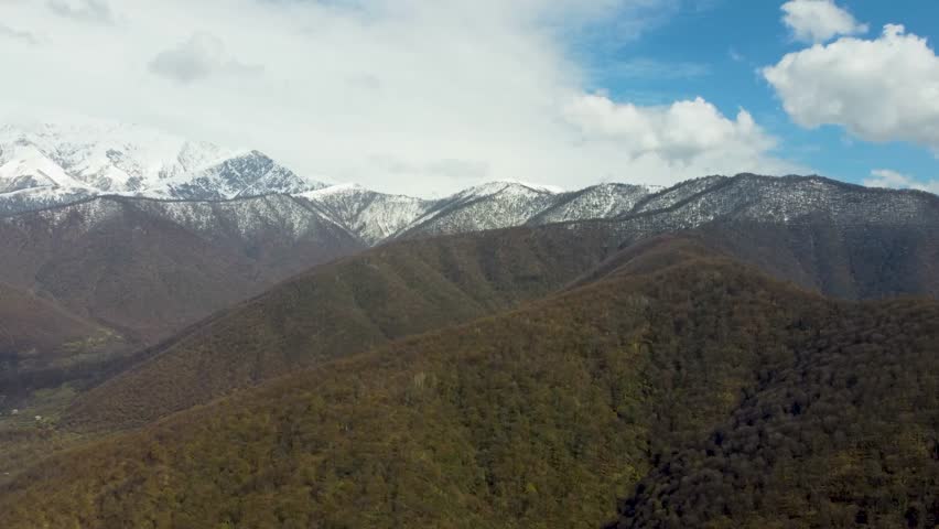 Snowy Mountain Peaks Behind Layered Forest Hills. Stunning aerial view showing the contrast between dark, wooded foothills and majestic snow capped mountains under a bright blue sky and fluffy clouds
