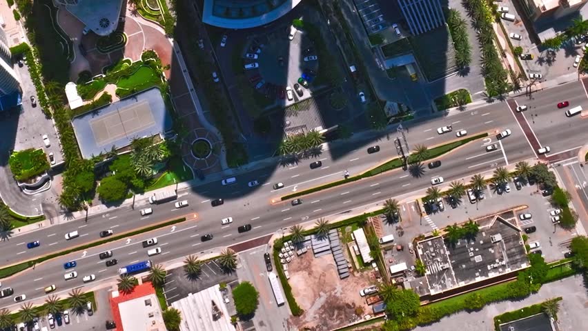 The scene shows an aerial view of busy streets in Miami. Cars move along the road while buildings and greenery are visible nearby. This captures city life in the daytime.