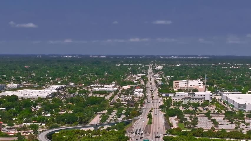 Streets and buildings in Miami during daytime. Cars travel along the road. Green trees and open areas are visible below, highlighting the urban landscape.