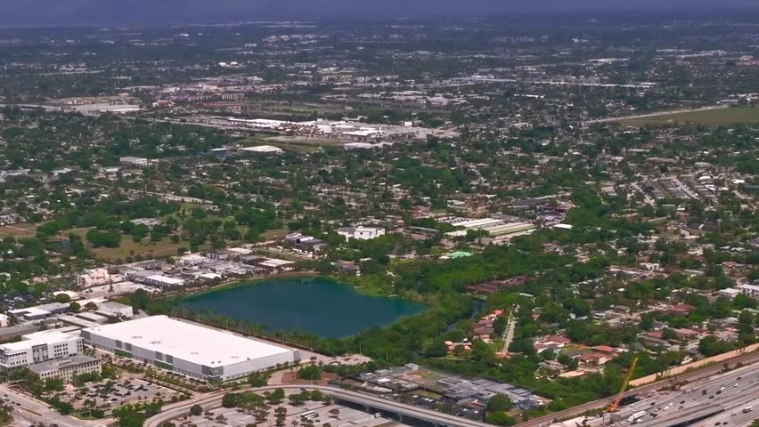 The image shows an aerial view of Miami. It features buildings, roads, green areas, and a lake. The landscape reveals the layout of the city and natural elements in the area.