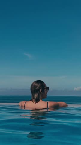 Relaxing Poolside Scene. Serene Woman Enjoying Bali Infinity Pool. Calm Woman Leaning On Edge Of Bali Infinity Pool. Peaceful Woman Relaxing At Infinity Pool With Sunglasses Reflecting Sky, vertical