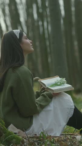 A young woman sits peacefully in a sun-dappled forest, engrossed in a book adorned with delicate white flowers, surrounded by trees and blooming ground cover