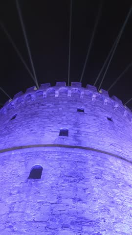 Vertical low angle shot looking up at the illuminated White Tower of Thessaloniki at night. Purple lighting and moving white searchlights against a dark sky, Macedonia, Greece.