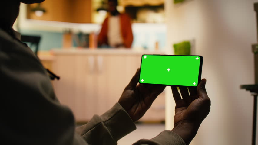 Isolated green screen on mobile phone next to black woman relaxing in hallway on the floor, browsing information and checking screen in calm domestic environment with soft light.