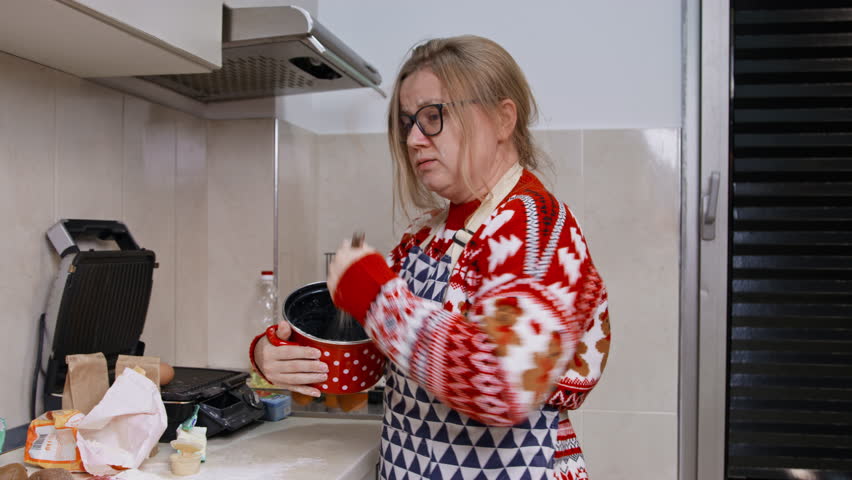 Adult woman with blonde hair in Christmas sweater whisking vigorously in bowl in kitchen with rushed stressed expression, chaotic cooking concept, busy homemaker scene, holiday kitchen humor.