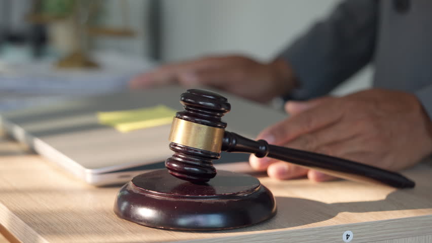 Wooden gavel rests beside open laptop. Hands type on keyboard, showing legal work. Gavel symbolizes judgment, law, and authority. Office desk setup suggests modern legal practice