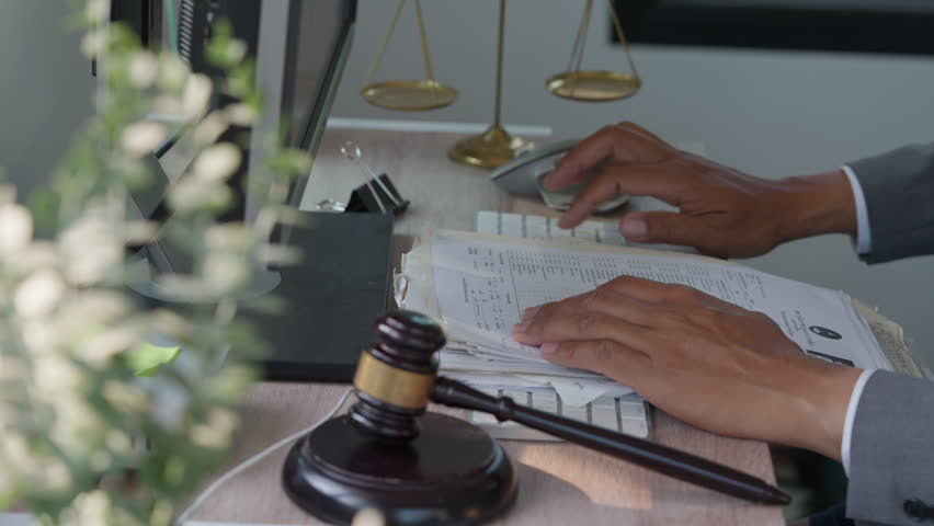 Lawyer reviews documents at desk. Gavel sits beside keyboard and mouse. Scales symbolize justice behind the hands. Green plant adds soft contrast to scene. Professional setting shows focused work