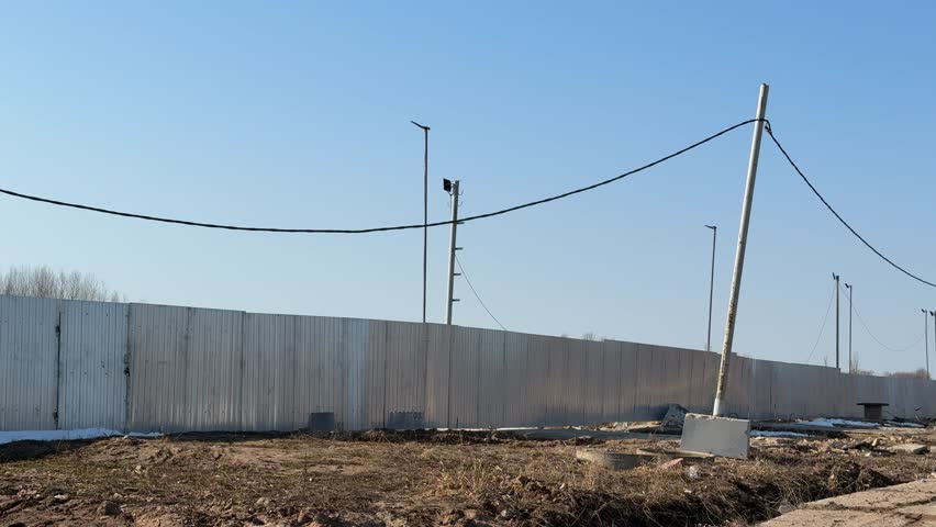 Construction site stainless steel fence against clear blue sky with lighting poles, utility wires, concrete bases and rough gravel.