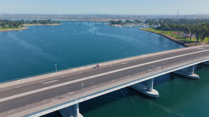 Drone footage capturing cyclists riding across concrete bridge over turquoise waters of Mission Bay in San Diego.