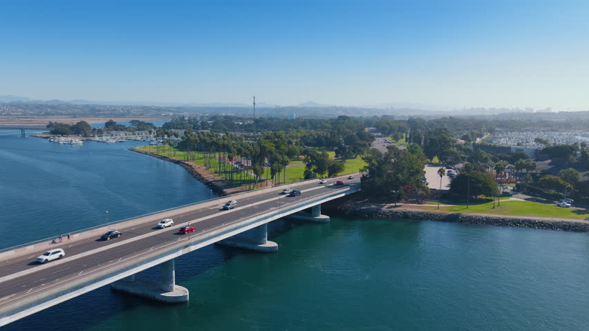 Drone footage of Mission Bay Park in San Diego showing cars crossing a bridge over turquoise waters. Palm trees line the green park area with marina and boats visible in the background on a sunny day