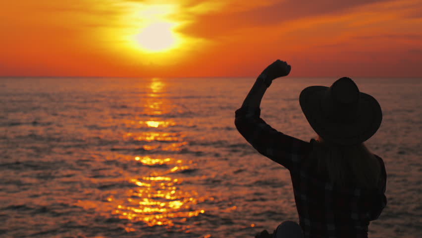Silhouette of young woman in cowboy hat sit on rocky cliffs at sunrise, raising hand in rebellious gesture, expressing freedom, attitude, and independent spirit.