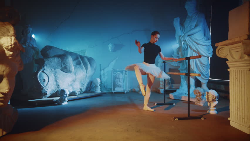 Beautiful woman in white tutu rehearsing in dark studio with statues, ballet. Classical art and dance performance in museum, graceful lady rising leg up, flexibility of human body, art performance