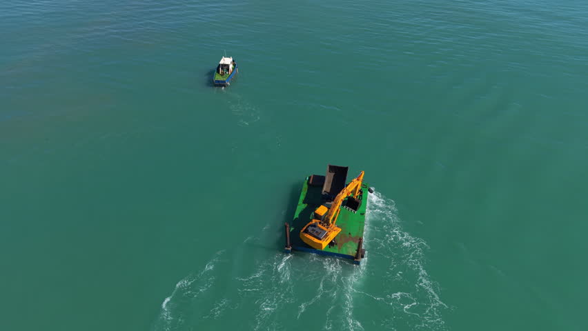 Overhead view of a yellow excavator mounted on a green barge operating in turquoise ocean water, with a small patrol boat nearby during marine construction work