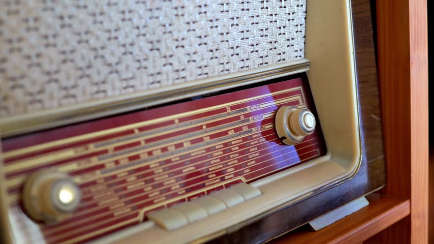 Close-up of a hand turning the tuning knob on a vintage radio receiver, showcasing the retro dial, wooden cabinet and textured speaker grille, evoking nostalgia and the analog listening experience
