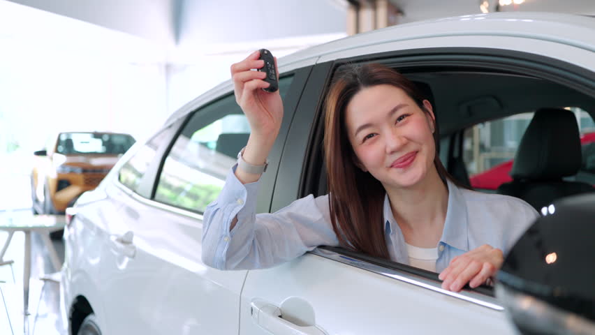 Happy young Asian woman smiling inside a new white car at an auto dealership showroom, proudly showing her new car key. Concept of buying a new vehicle.