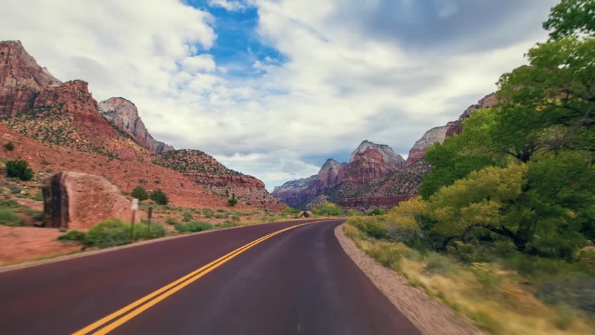 wide-angle view from a moving vehicle captures the majestic landscape of Zion National Park in Utah. The winding road leads into a canyon surrounded by towering, reddish-orange sandstone cliffs and ro