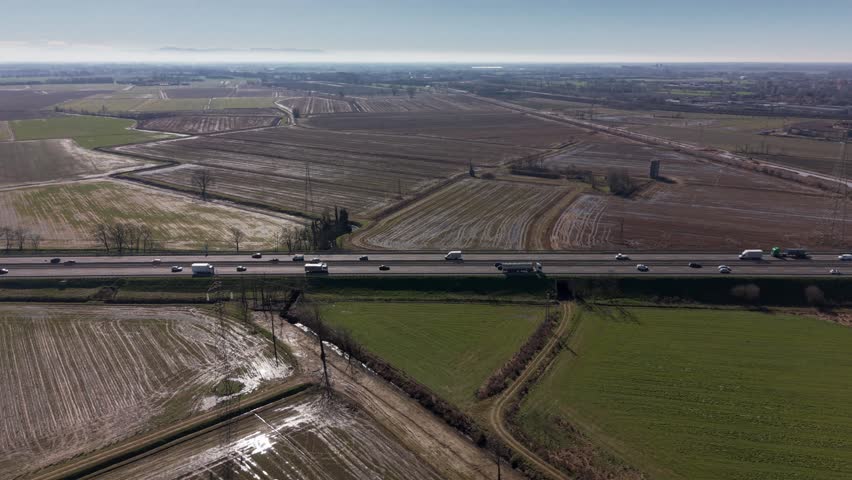 Elevated perspective of cars and trucks driving on a busy highway that cuts through vast agricultural fields and countryside landscape