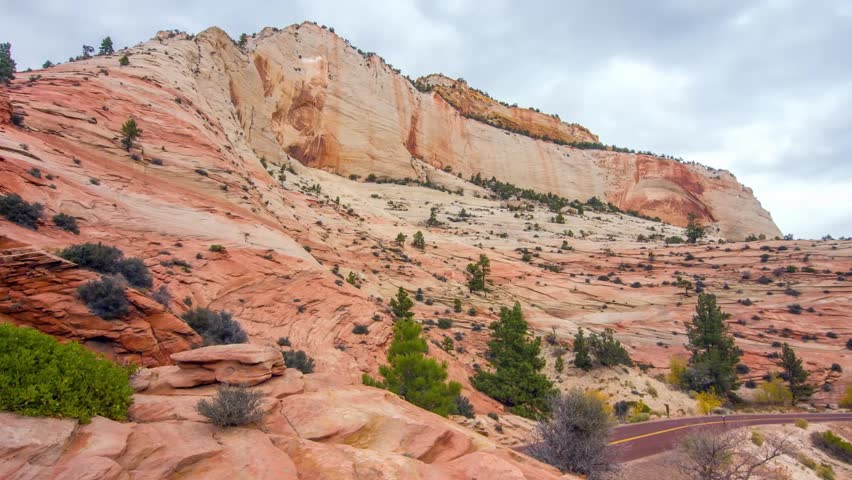 A wide shot of the stunning scenery in Zion National Park, Utah, featuring immense towering red and white sandstone cliffs with unique swirling strata and sparse evergreen vegetation under an overcast