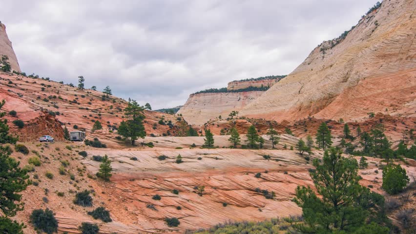 A wide, scenic view of the striking red rock formations and sandstone mountains within Zion National Park, Utah, USA. The landscape features a winding road, sparse green trees and shrubs, and a dramat