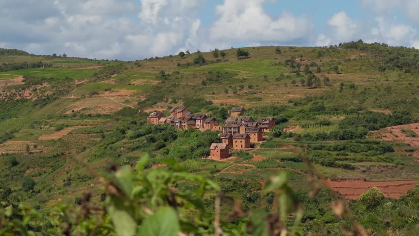 A scenic wide shot showcasing a traditional village with brick houses nestled on a lush green hillside under a blue cloudy sky in the rural highlands of Madagascar AfricaVillage, Madagascar, Highlands