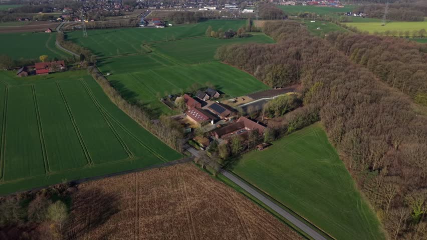 Aerial flyover of rural countryside farmstead surrounded by green fields and forest in spring, featuring residential buildings with solar panels, farmland patterns and peaceful natural landscape.