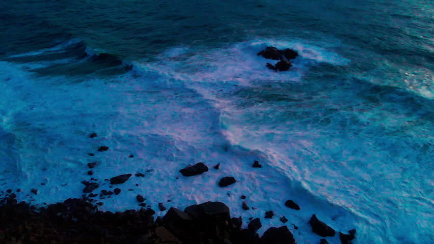 Drone flying over ocean waves and rocks in blue early sunrise light with dramatic sea texture