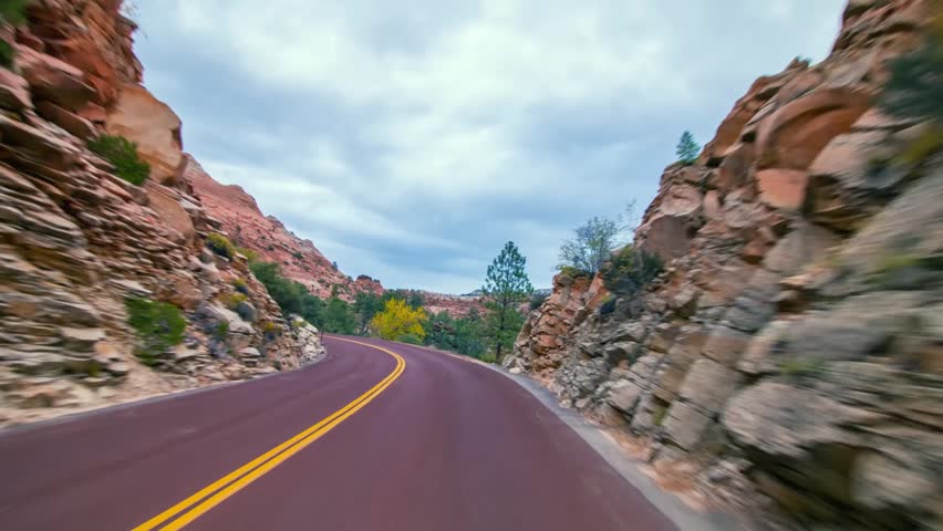 Scenic footage from a moving vehicle captures the beautiful landscape of the Zion-Mount Carmel Highway in Zion National Park, Utah. Towering reddish sandstone cliffs and sparse green vegetation line t