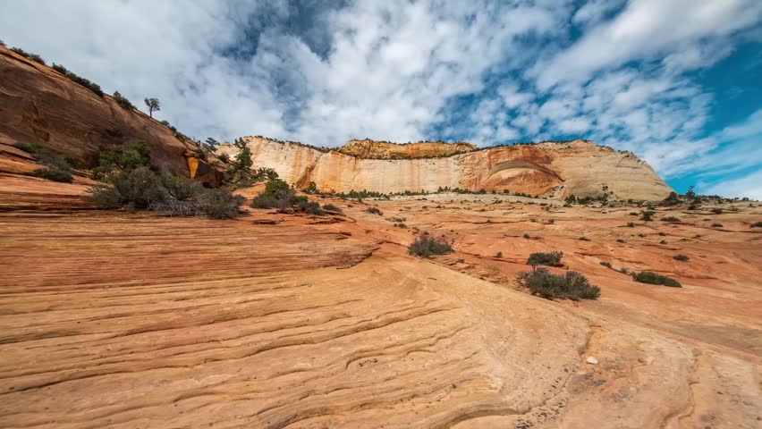 A wide-angle shot capturing the stunning, rugged landscape of Zion National Park in Utah, USA. The foreground features textured, wavy red rock formations, leading the eye towards massive, towering san