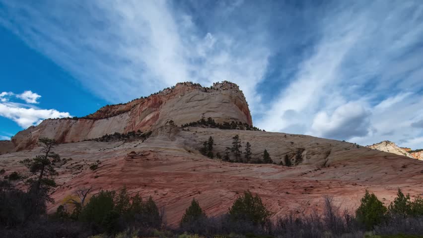 An establishing shot of massive, layered red and white sandstone formations under a bright blue sky with dynamic white clouds in Zion National Park, Utah, USA.
