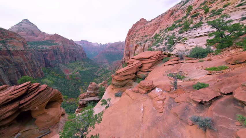 A high-angle panoramic view of the majestic Zion Canyon at the end of the popular Canyon Overlook Trail. Towering red and orange sandstone cliffs and dramatic rock formations dominate the landscape, w