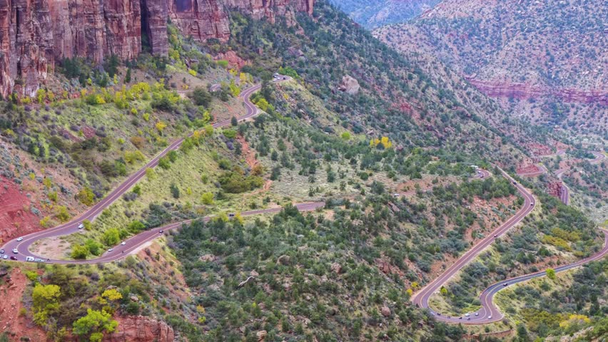 A high-angle, scenic view of the winding Zion-Mount Carmel Highway as it makes its way through the steep, red sandstone cliffs and canyons of Zion National Park in Utah. The dramatic hairpin turns are