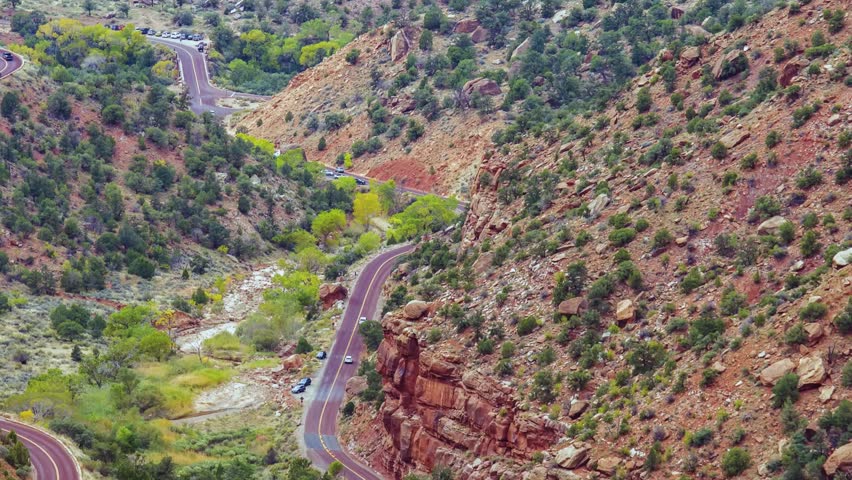 A high-angle, wide-shot view of the winding Zion-Mount Carmel Highway as it cuts through the dramatic, rugged red-rock landscape of Zion National Park, Utah.
