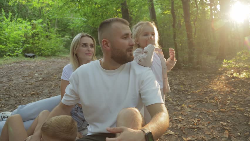 Warm family moments unfolding during woodland picnic, golden sunlight streaming through trees while parents and children sharing joyful connection