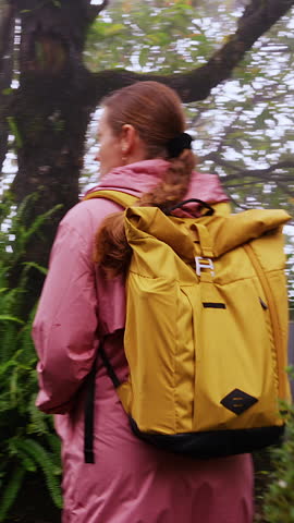 Vertical video. Woman standing on misty Forest Path with yellow Backpack in a Wide Angle view showing ferns, trees, wet stones and soft atmospheric light.