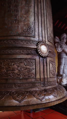 Vertical video. Massive Decorated Bell With Wooden Striker Ready to Hit Surrounded by Carved Statues in Low Angle Wide View at Ba Na Hills Vietnam, Spiritual Tradition.