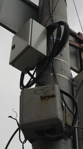 Close-up view of weathered electrical junction enclosures mounted on a utility pole with cables, conduit, and straps, highlighting corrosion and aging hardware in infrastructure