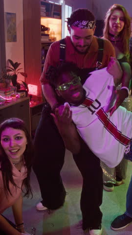 Vertical dolly in portrait of diverse young adults lifting friend in arms and cheering while posing and gesturing toward camera at house party