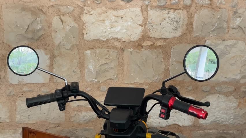 Motorcycle handlebars against a rustic stone wall background, featuring mirrors and controls video