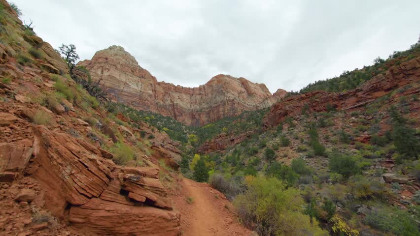 A high-angle view looking down at the red rock formations and desert greenery along the Watchman Trail in Zion National Park, Utah, under a cloudy sky.