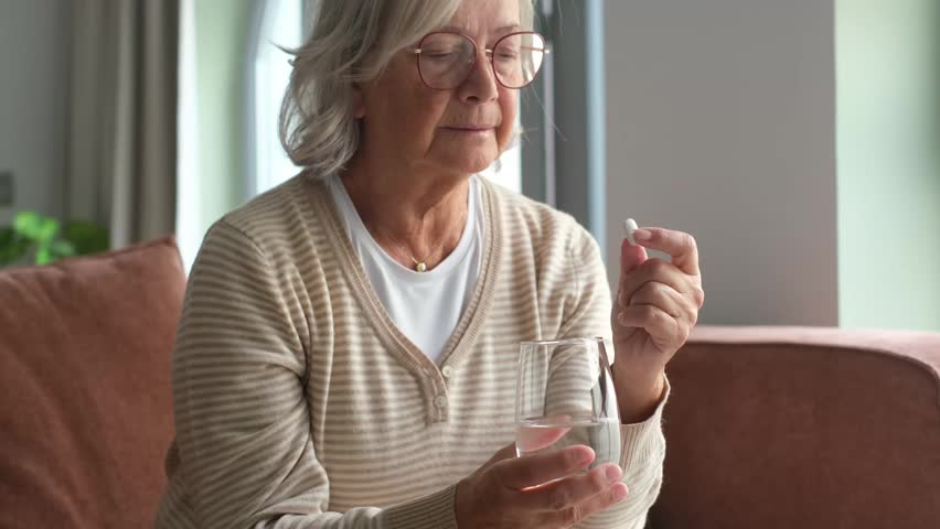 An elderly woman sits calmly on a sofa, holding a white pill and a glass of water. She prepares to take her daily medication, highlighting themes of health, wellness, and self-care in old age.