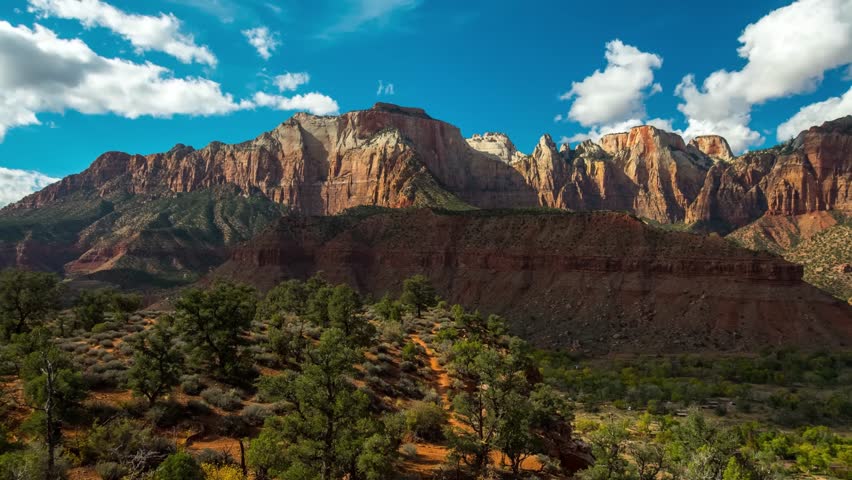 A scenic view from the moderately difficult Watchman Trail in Zion National Park, Utah, capturing the dramatic red rock formations and cliffs under a blue sky with white clouds.