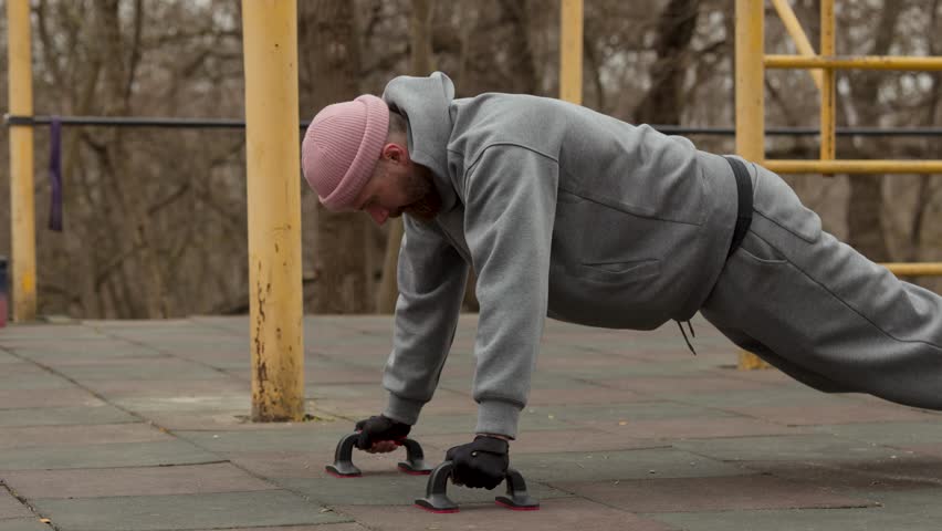 Side view of male athlete performing push ups using floor bars on outdoor sports ground. Bearded man in pink beanie training chest and arms during morning session
