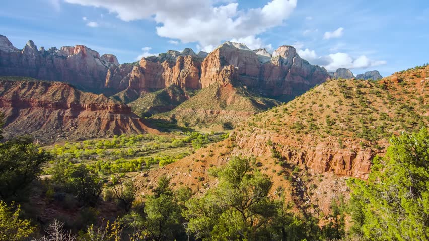 A panoramic view of the rugged, reddish-tan Navajo sandstone cliffs and the lush green canyon floor of Zion National Park, Utah. The footage captures the iconic Watchman peak towering over the landsca