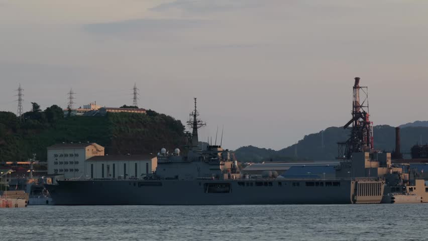 Japan Maritime Self-Defense Force (JMSDF) transport ship docked at Kure Port at dusk. An industrial maritime landscape featuring naval vessels and port facilities under an evening sky.